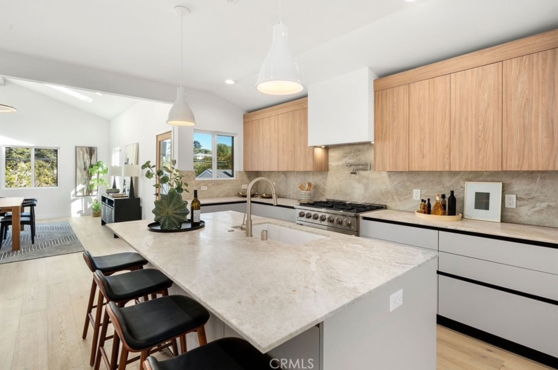 Modern kitchen with a large stone island, light wood upper cabinets, gray lower cabinets, and black bar stools.