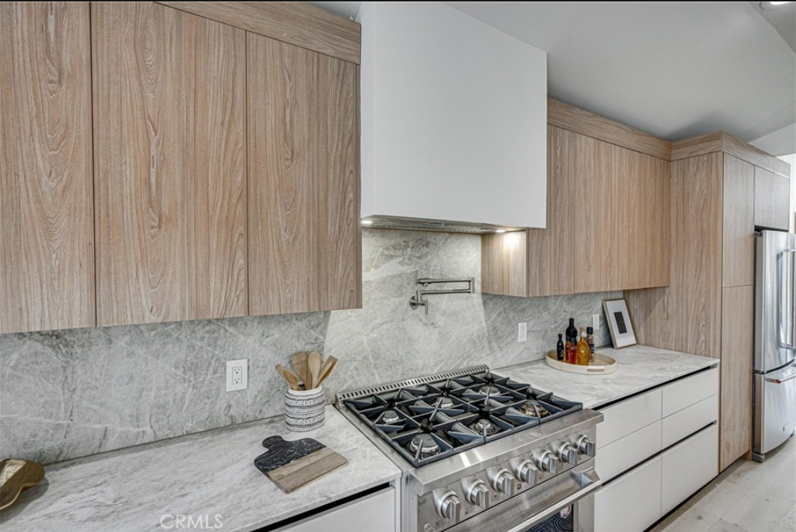 A modern kitchen features light wood cabinets, a white range hood, a marble backsplash and countertops, and a gas stove.