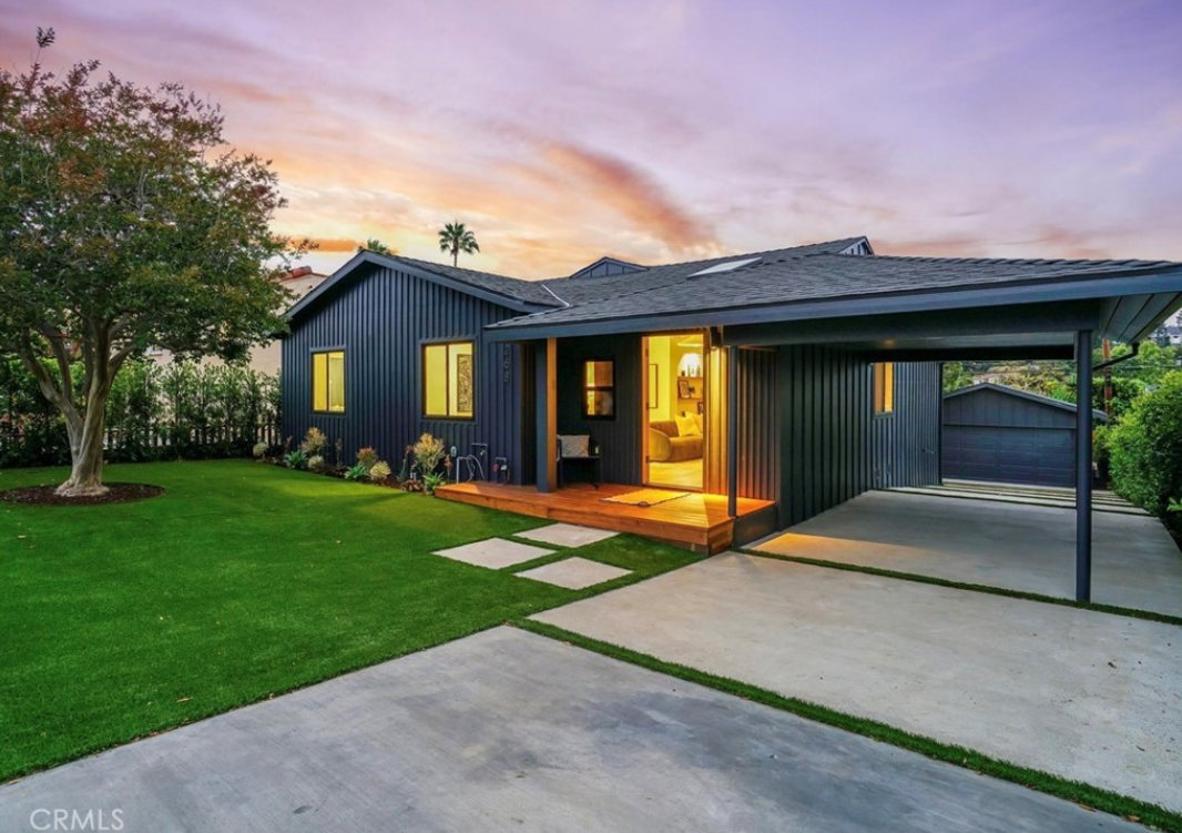 A modern, dark-sided single-story home with a wooden porch and attached carport, set against a sunset sky.
