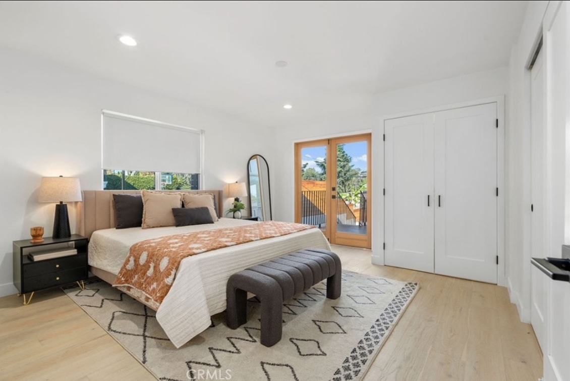 Bright bedroom with a king-sized bed, light wood floors, a patterned rug, white walls, and a glass-paneled door.