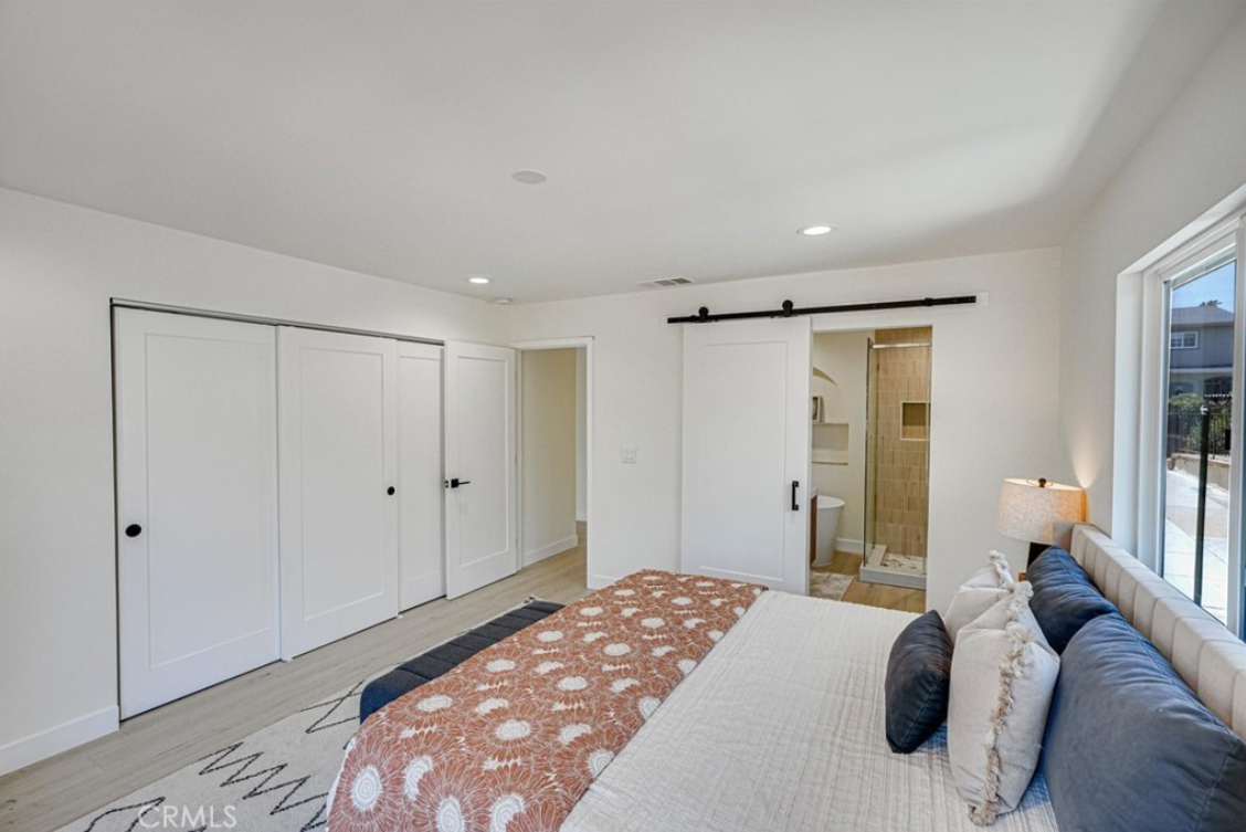 A modern bedroom with a large bed, white closet doors, a sliding barn door leading to a bathroom, and a bright window.