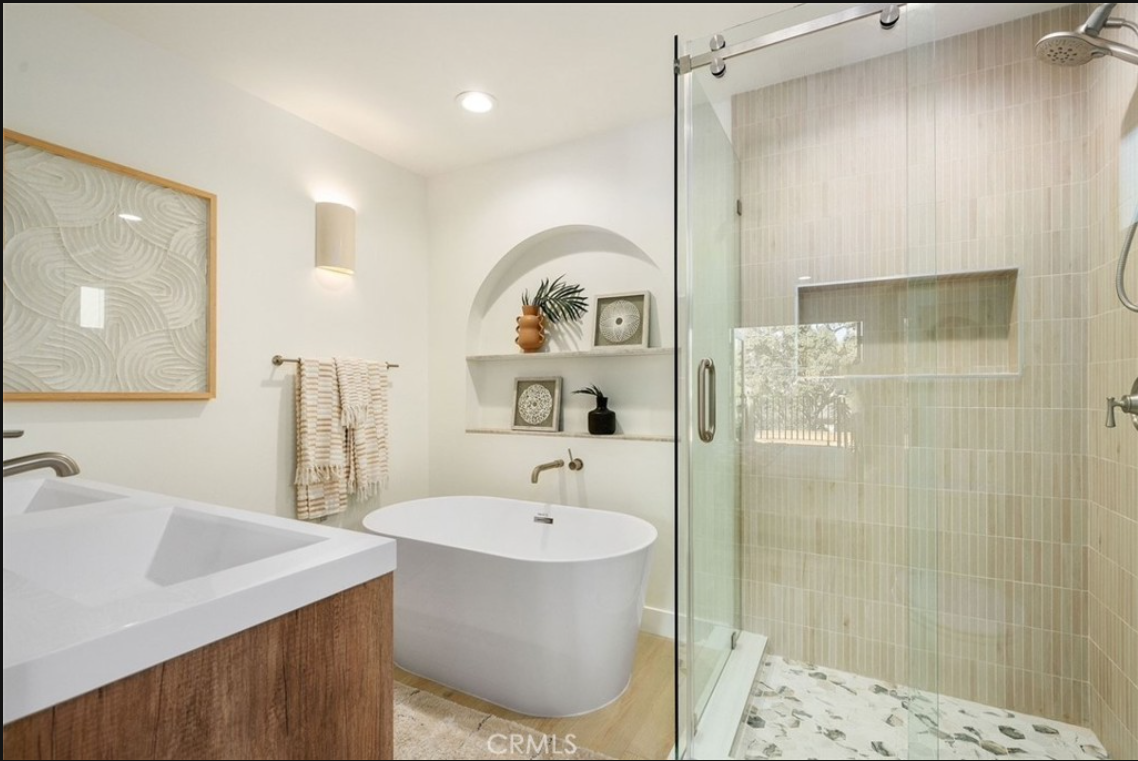 A modern bathroom featuring a white freestanding tub, arched shelving, wood-tone vanity, and a glass-enclosed shower.