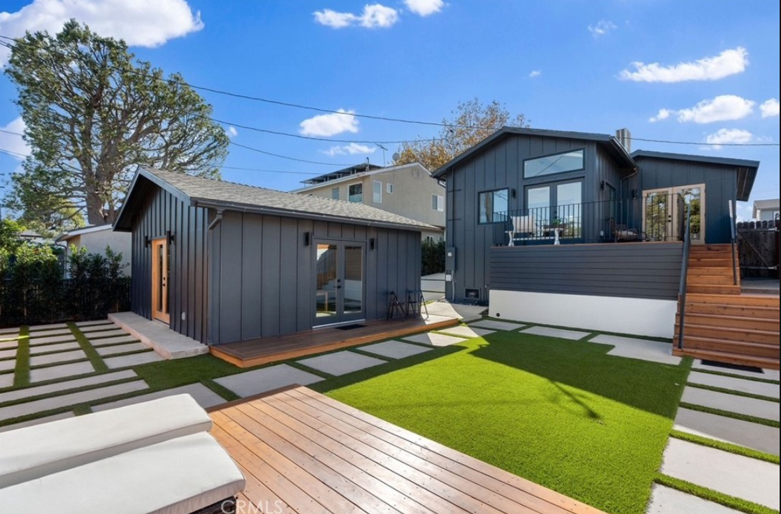 Modern backyard with a dark-sided structure, wooden deck, artificial lawn, and paved stone pathway under a blue sky.