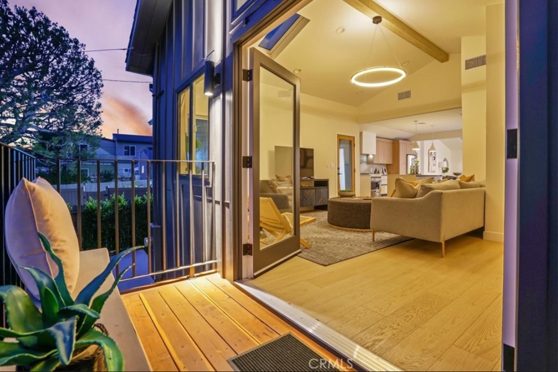 A sunset view from a balcony looking into a modern living room with light wood floors and a beige sofa.