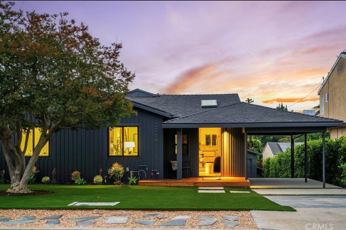 A modern single-story house with dark vertical siding, a wooden front porch, and a carport at sunset.
