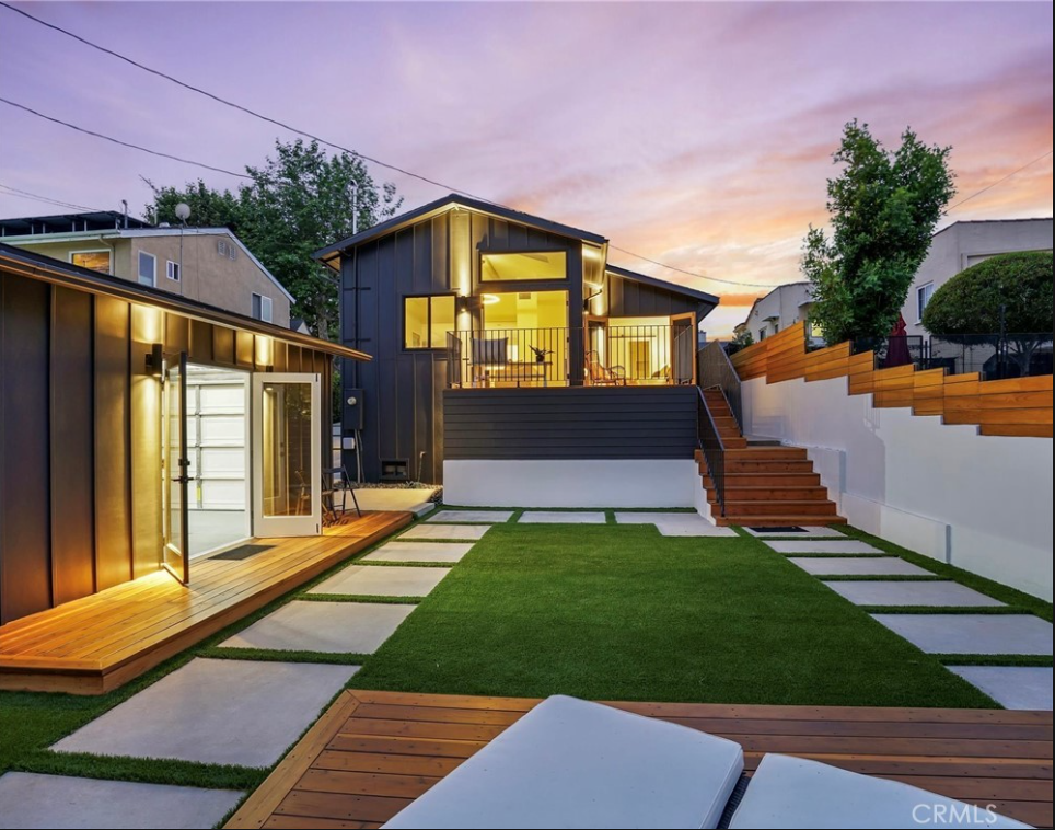 A backyard at sunset featuring a modern house, a matching studio, a lush green lawn, and tiered wooden steps.