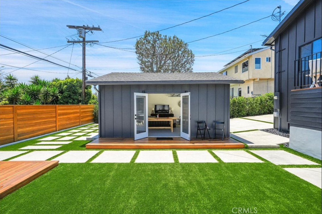 A gray detached studio with open French doors, a wooden deck, and a yard with pavers and artificial grass.