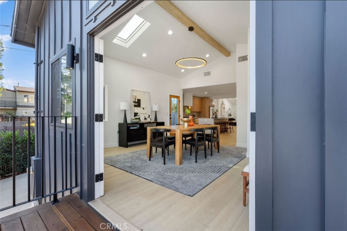 A view from a patio into an open dining room with light wood floors, a wooden dining set, and a geometric area rug.