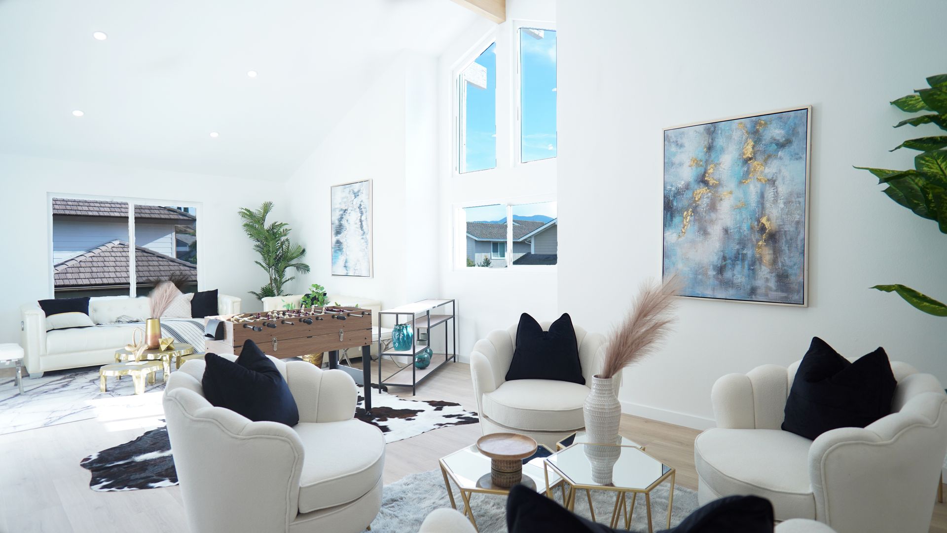 A bright, modern living room with white walls, cream armchairs, a foosball table, abstract art, and large windows.