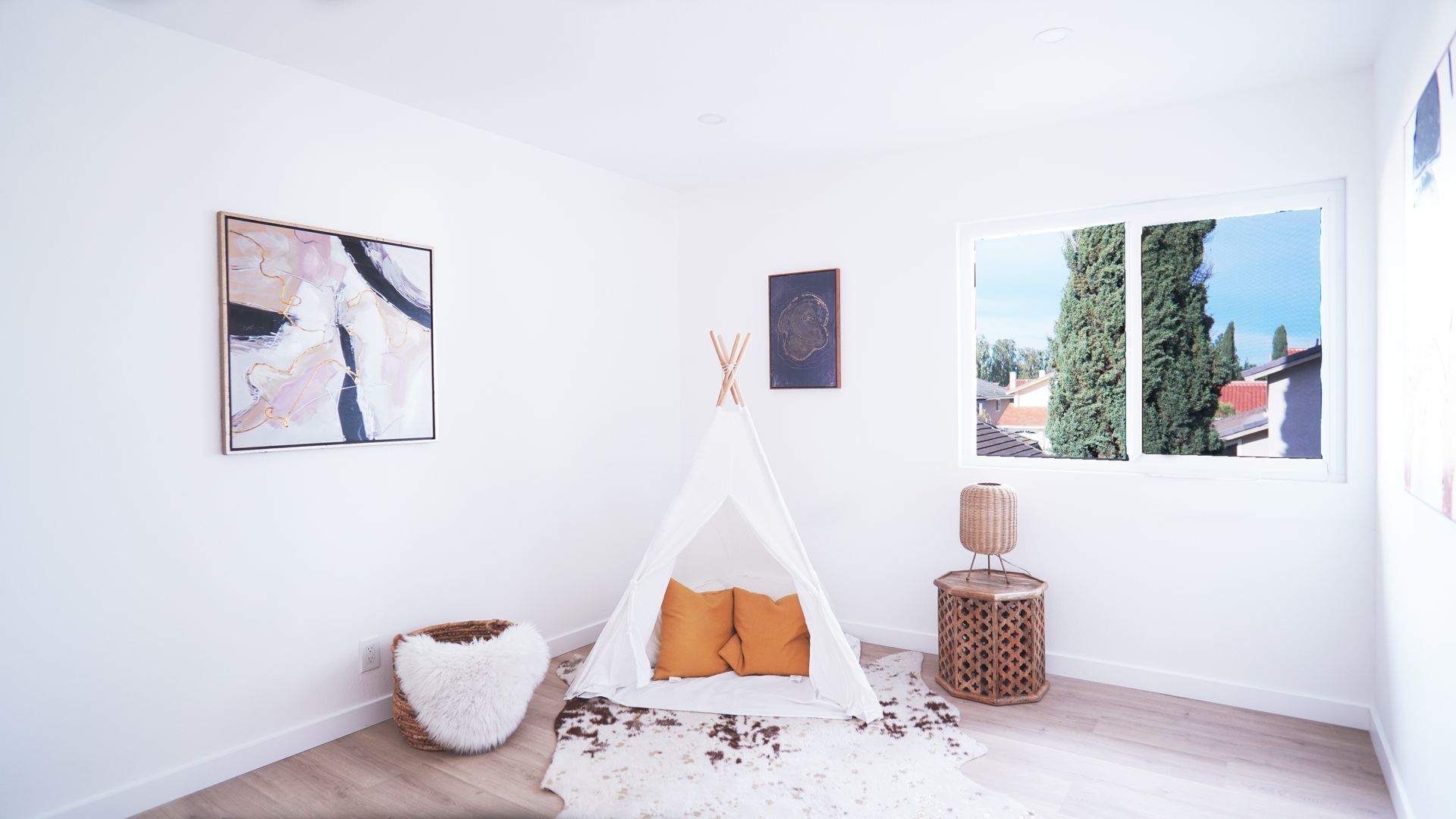A bright, minimalist room features a white play tent with mustard pillows, a faux fur rug, and wicker furniture.