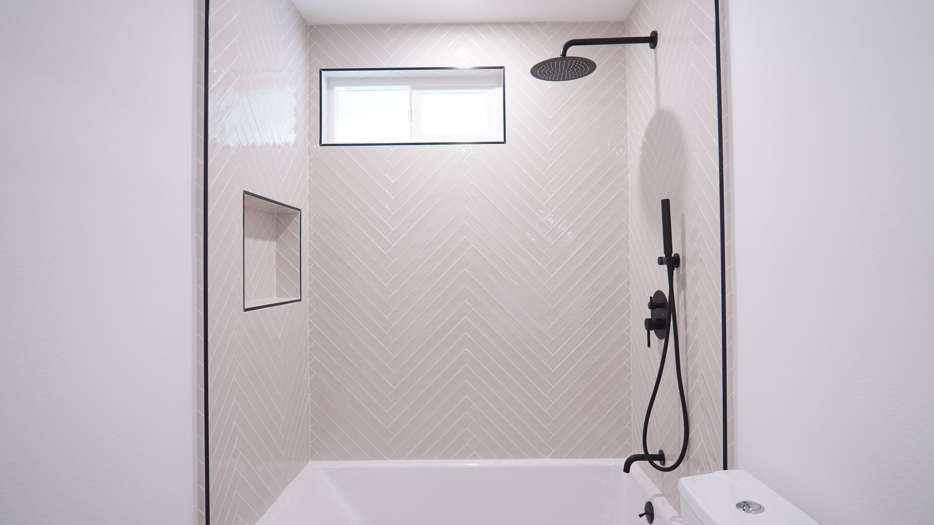 A shower-tub combo with light-colored herringbone wall tiles, black fixtures, and a recessed wall niche.