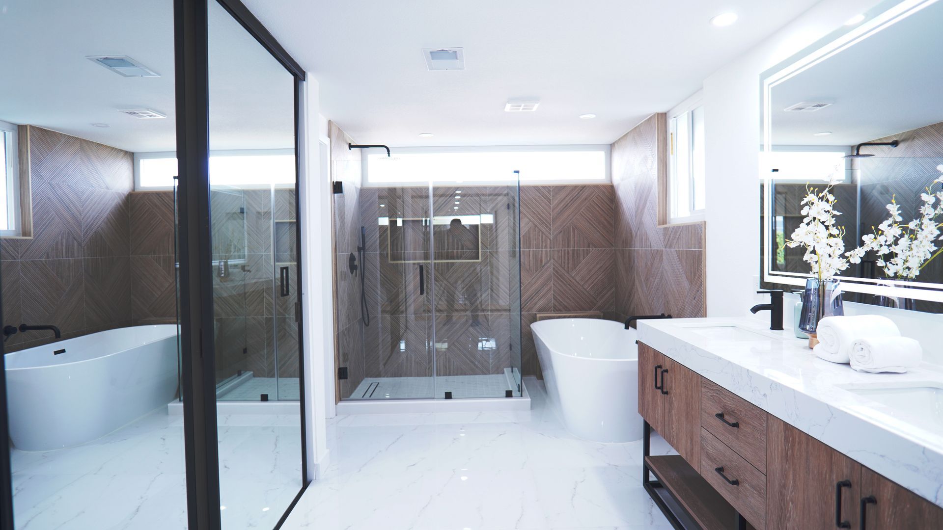 A modern bathroom with a double vanity, a glass-enclosed shower, a freestanding tub, and brown wood-patterned accent walls.