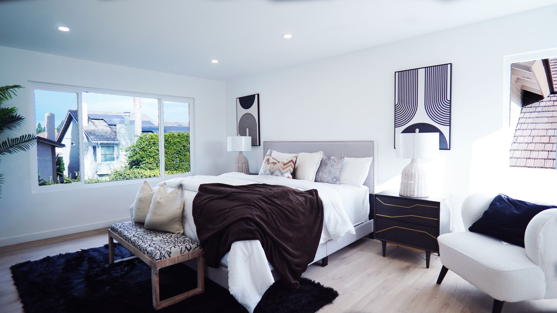 Modern bedroom with a gray bed, white bedding, dark throw blanket, dark area rug, bedside tables, and a lounge chair.