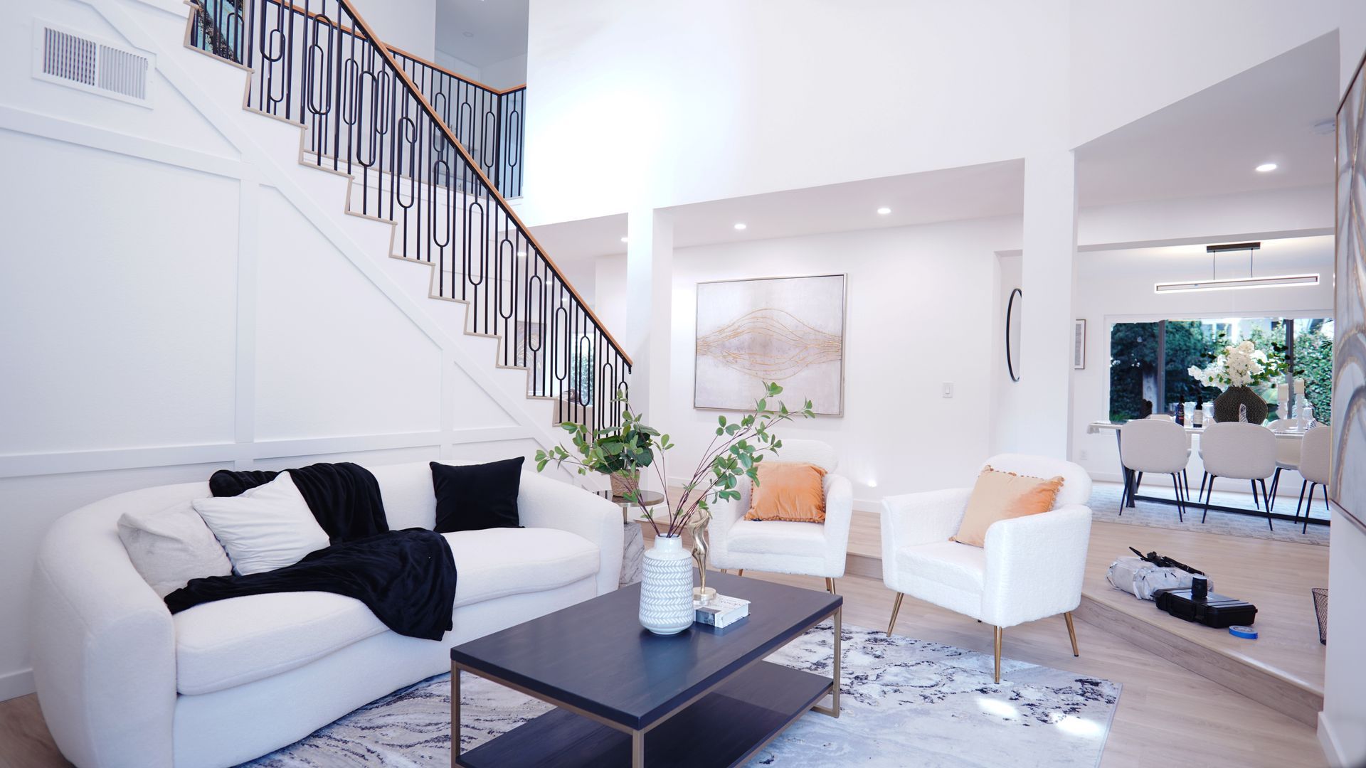 A bright, modern living room with white sofas, a dark coffee table, and an open dining area, featuring a staircase.
