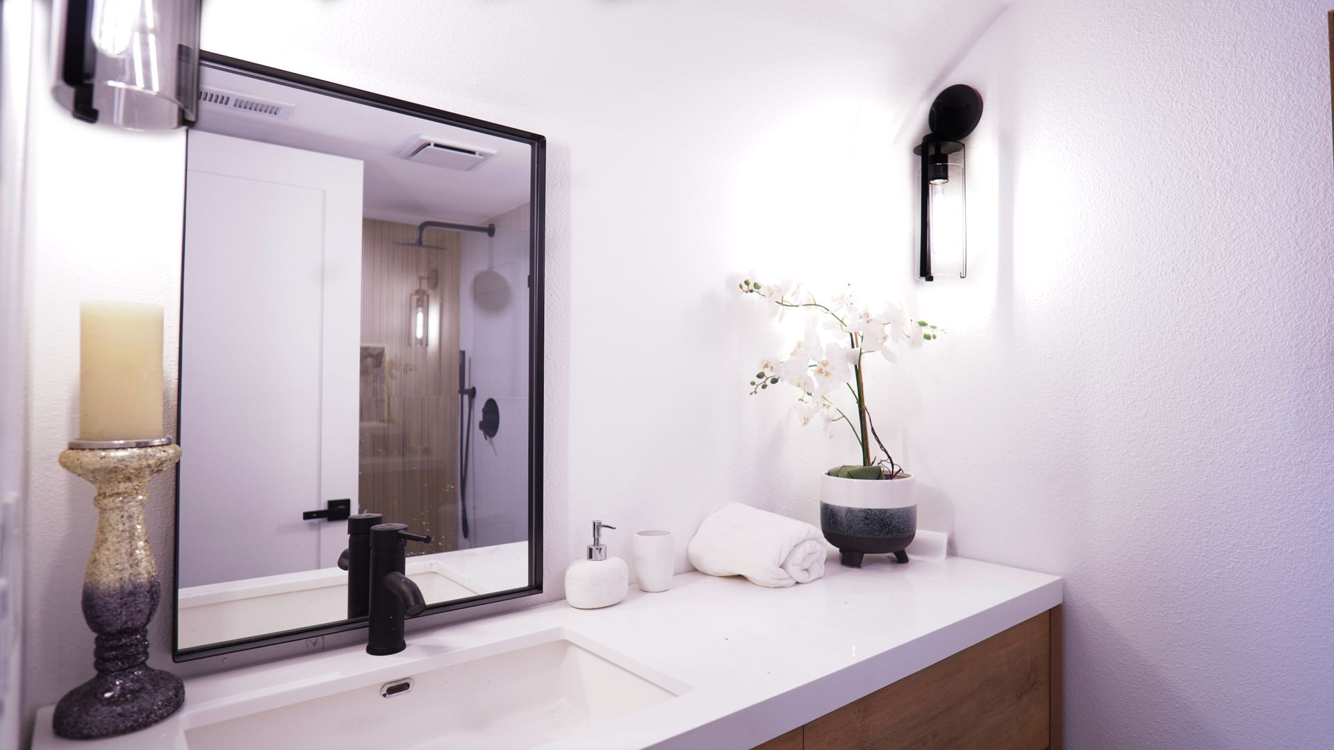 Modern bathroom vanity with a white countertop, sink, black-framed mirror, candle, potted orchid, and a white towel.