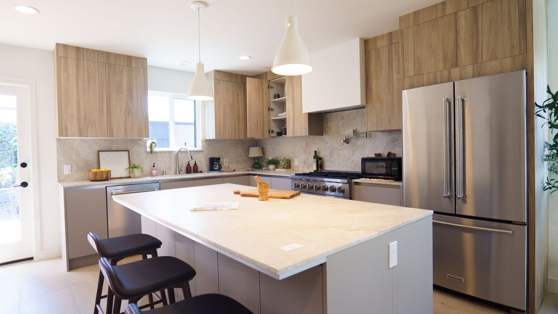 Modern kitchen featuring a large white island with three stools, stainless steel appliances, and wood-toned cabinetry.