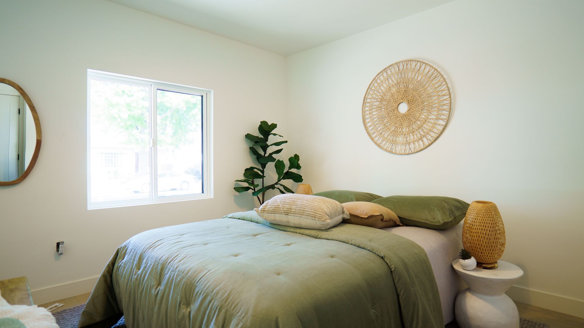 A bedroom with a green comforter, a white bedside table, a potted plant, and a large round woven wall art piece.