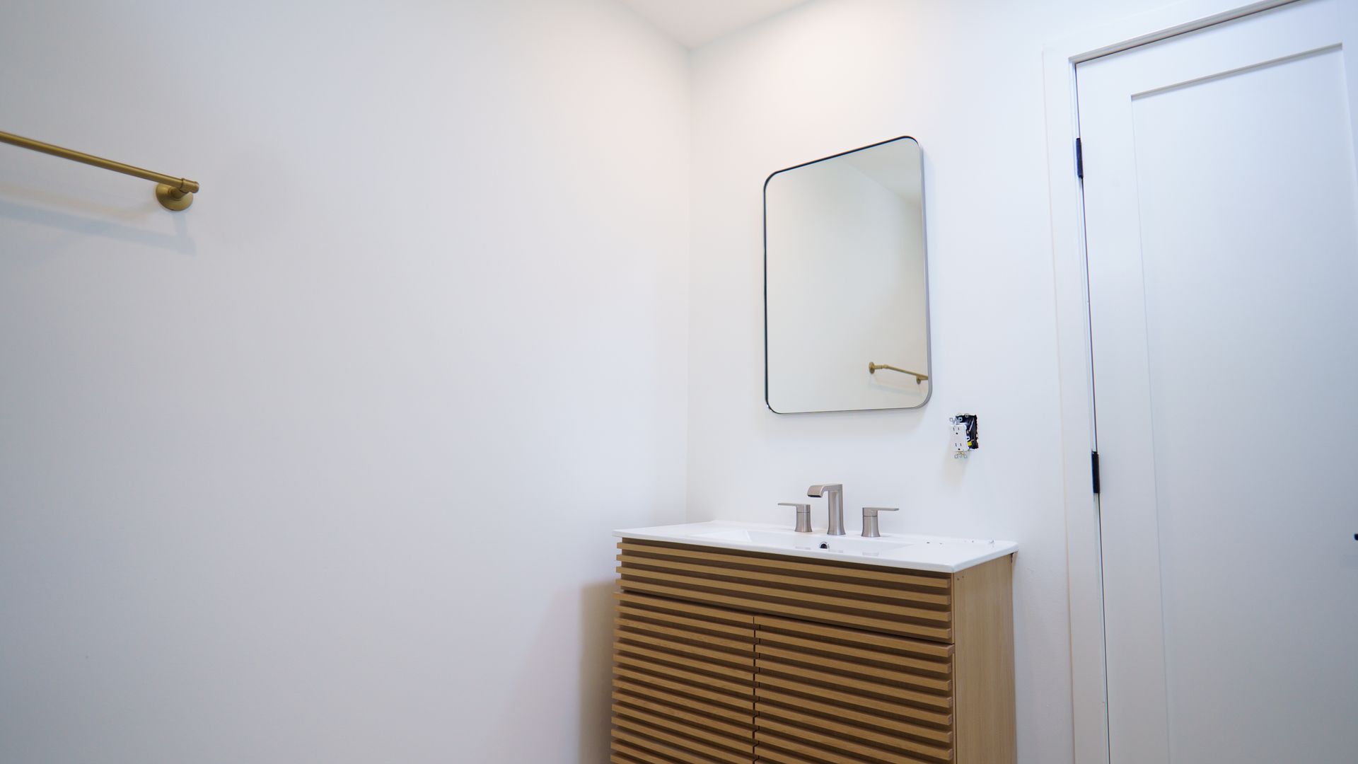 A modern bathroom featuring a white wall, a brass towel bar, a rectangular mirror, and a wood-textured vanity with a sink.