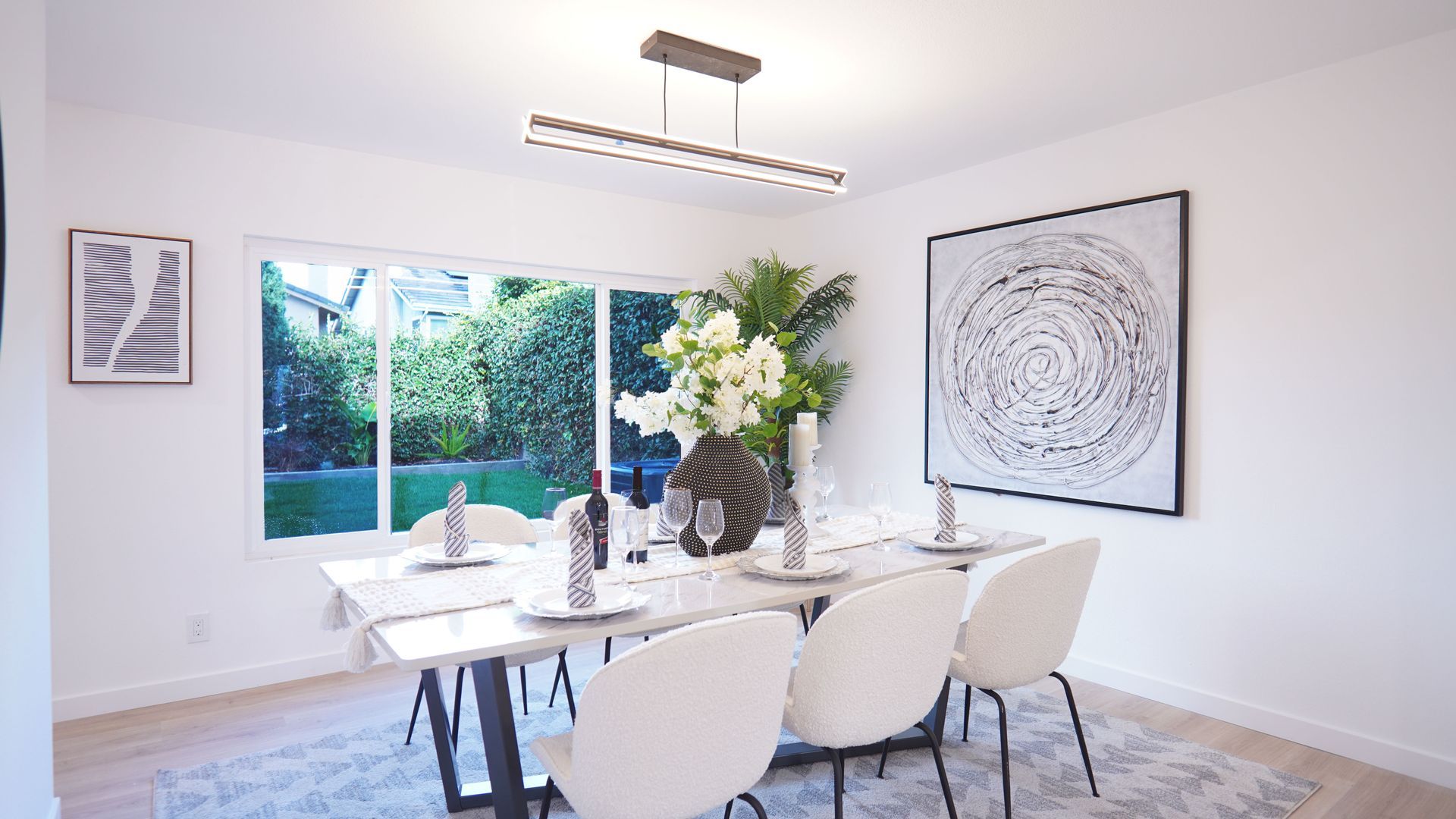A bright dining room with a white table, modern white chairs, a pendant light, and large wall art, overlooking a garden.