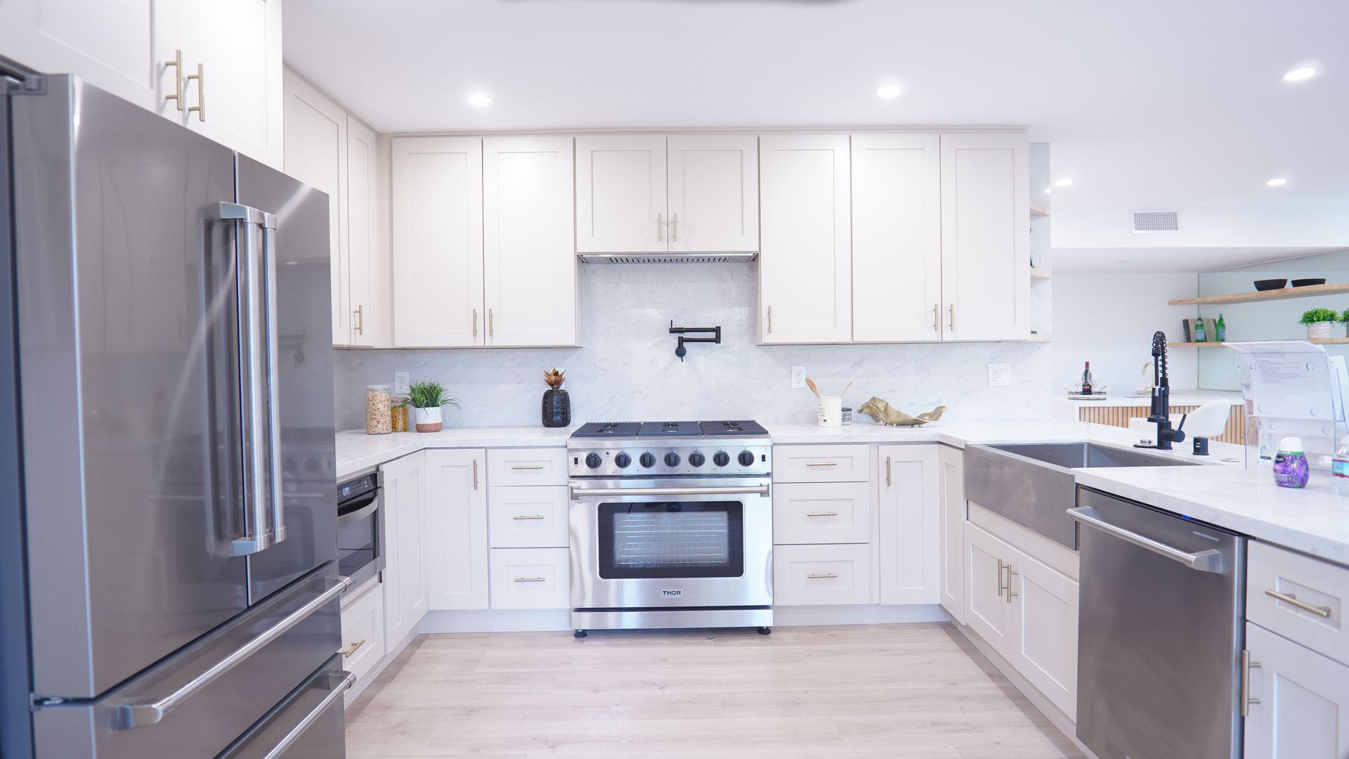 A modern kitchen with white shaker cabinets, light wood flooring, stainless steel appliances, and a farmhouse sink.