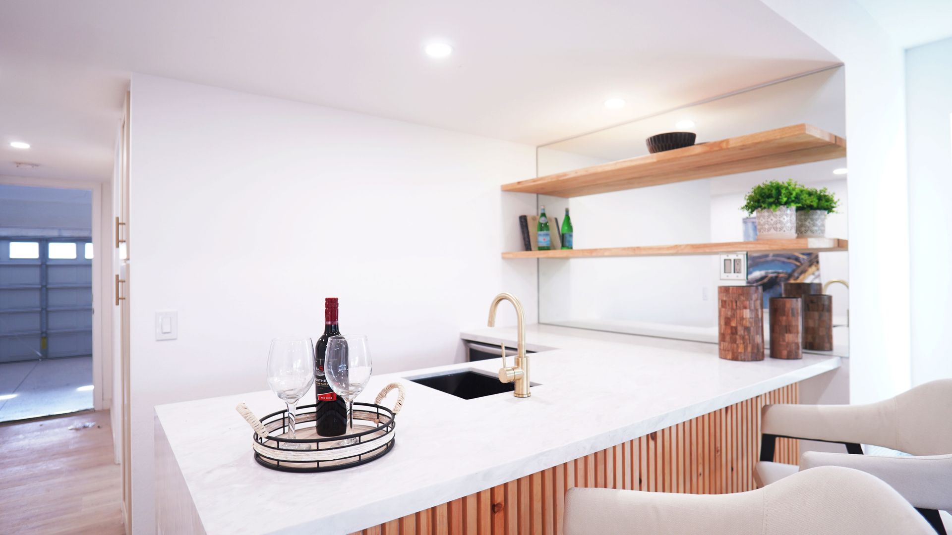 A modern kitchen island with a wine bottle and glasses, featuring light wood slat cabinetry, white counters, and shelving.