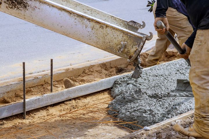 Construction workers use a chute to pour wet concrete into a framed-out form on a dirt construction site.