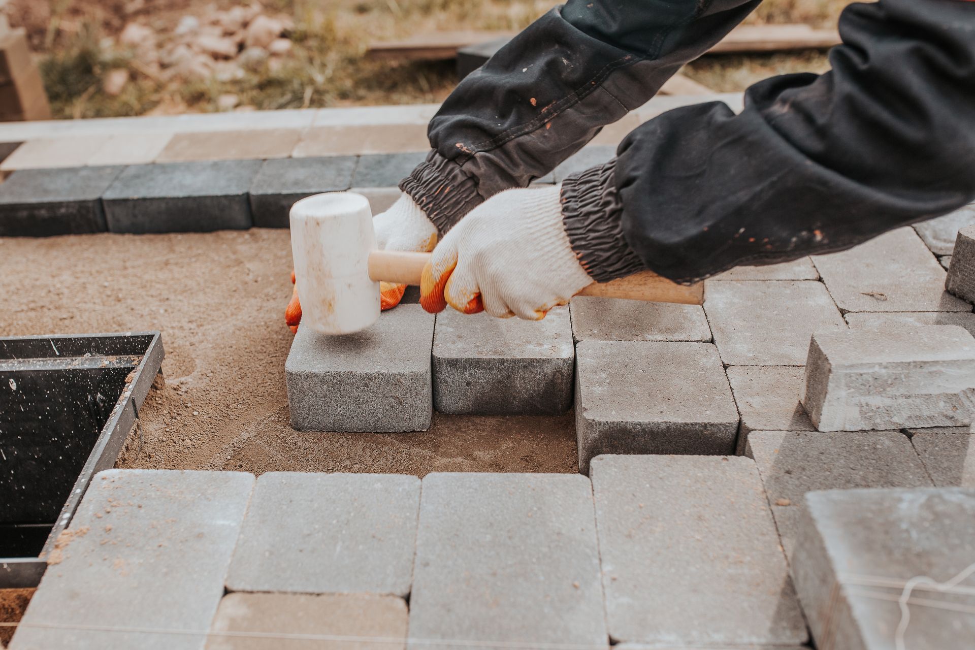 A person in work gloves uses a white rubber mallet to set gray paving stones into a sand base.