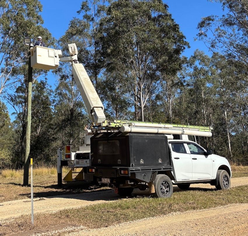 A utility worker in a cherry picker bucket, working on a utility pole. White truck and green trees in background— Gold City Solar & Electrical in Long Flat, QLD