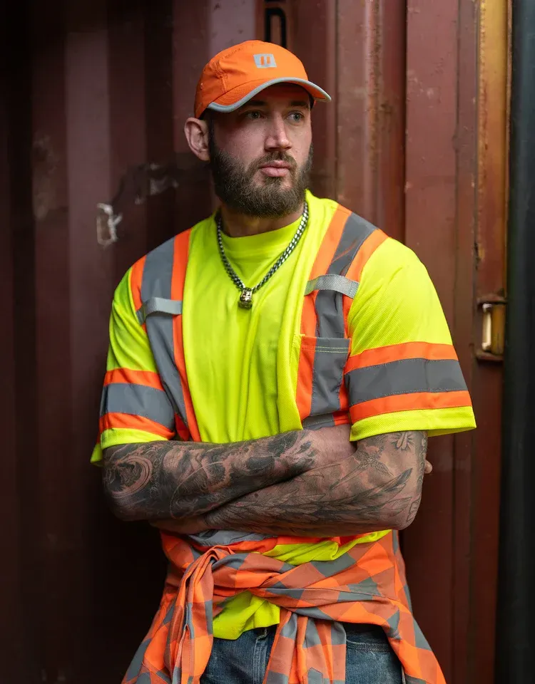 Man in neon safety gear leans against a brown shipping container, arms crossed, looking away.