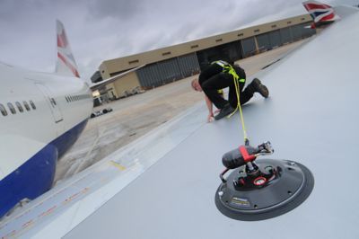 Person on airplane wing using a suction device for maintenance. Aircraft in background, overcast day.