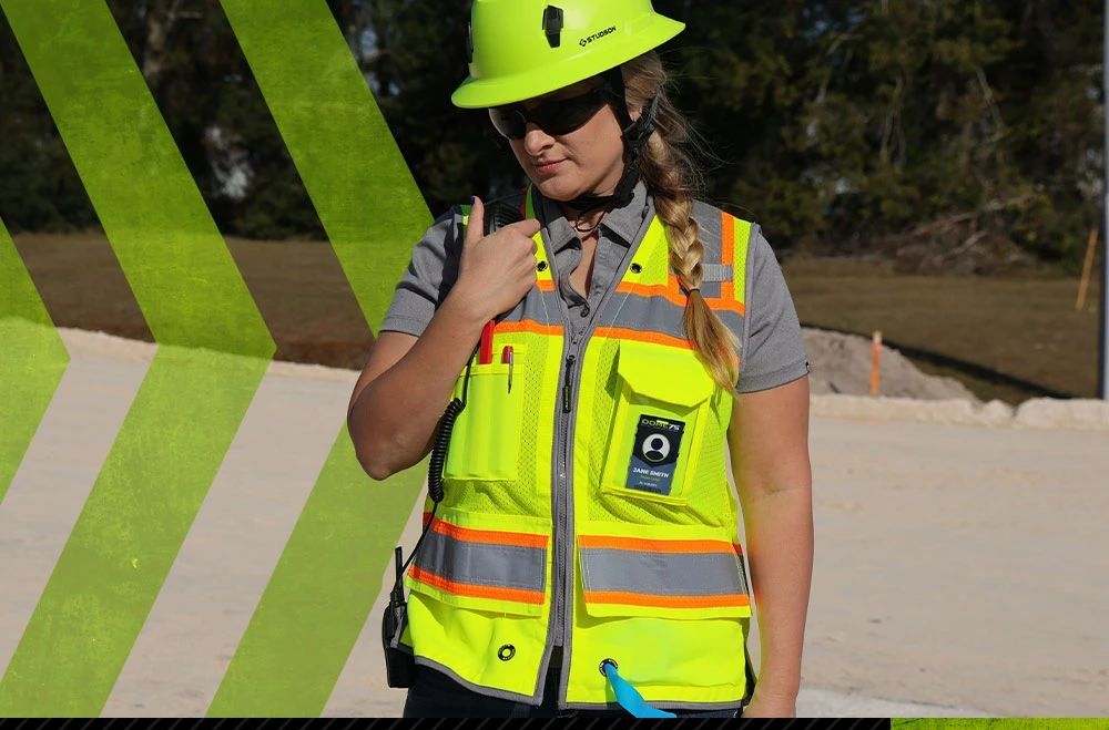 Man in neon safety gear leans against a brown shipping container, arms crossed, looking away.