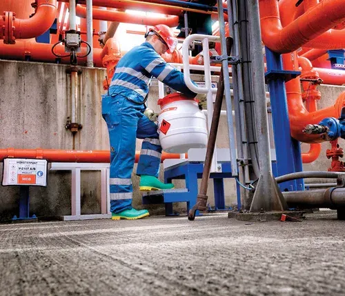 Worker in blue coveralls and hard hat tending to a chemical container near red pipes.