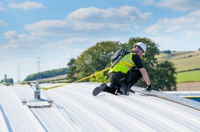 Worker in safety gear on a metal roof, attached to a rope, under a blue sky.