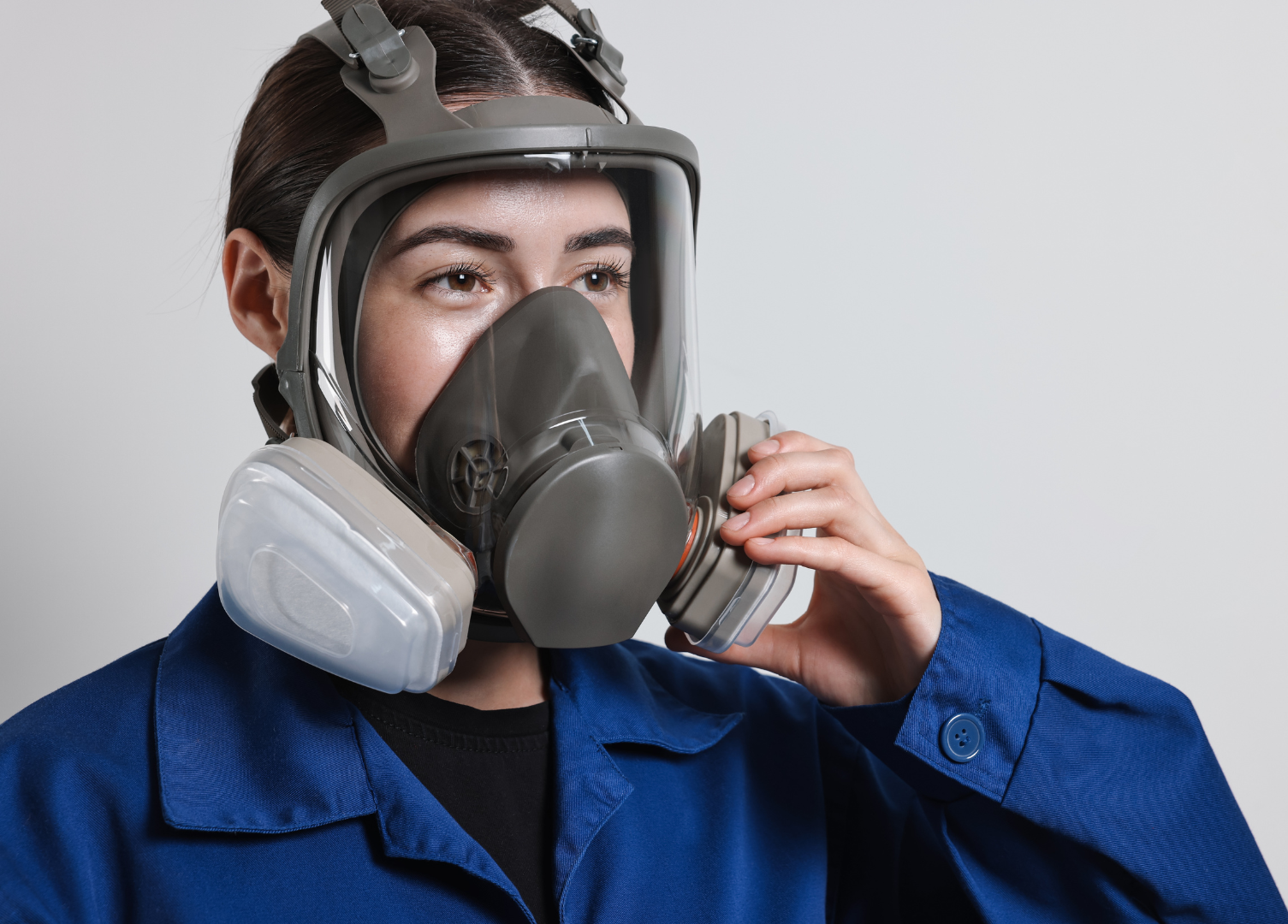 Woman in blue work clothes adjusting a respirator mask with white filters.