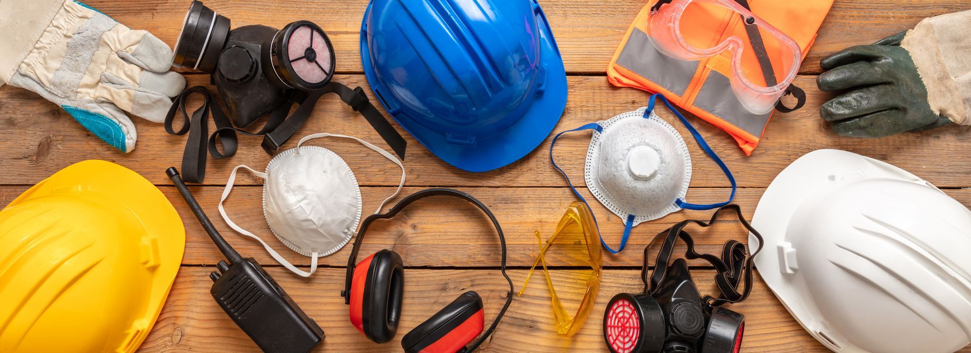 Safety equipment, including hard hats, respirators, safety glasses, gloves, and ear protection, arranged on wood.