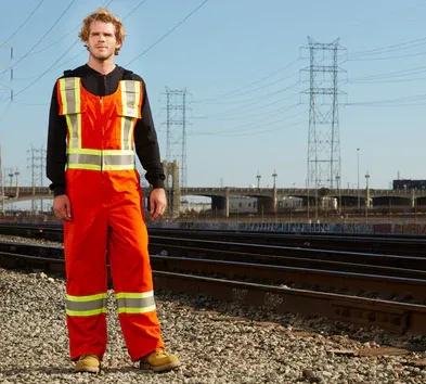Man in orange work coveralls stands on gravel next to train tracks, power lines in background.