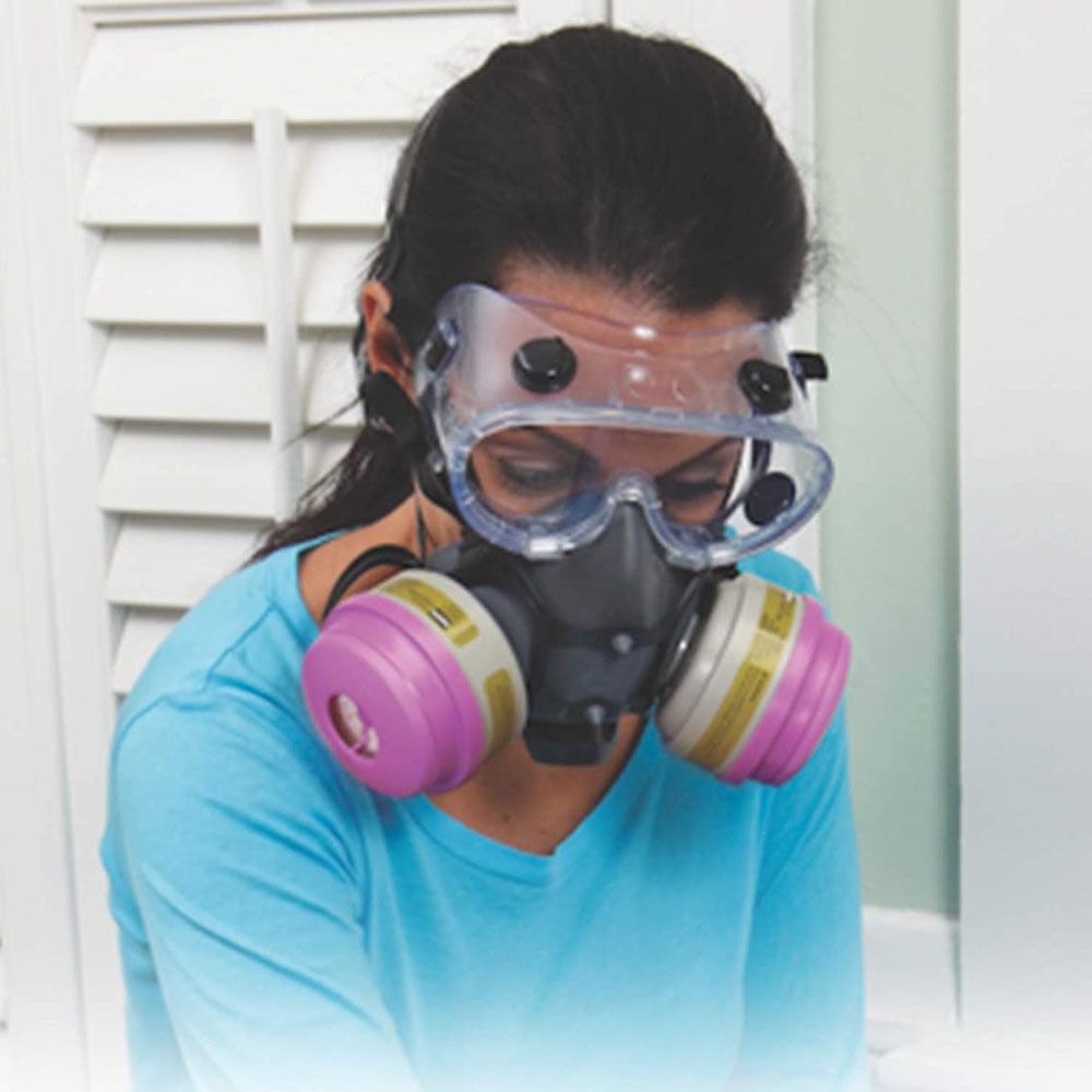 Woman wearing safety goggles and respirator mask indoors, near a shuttered window.