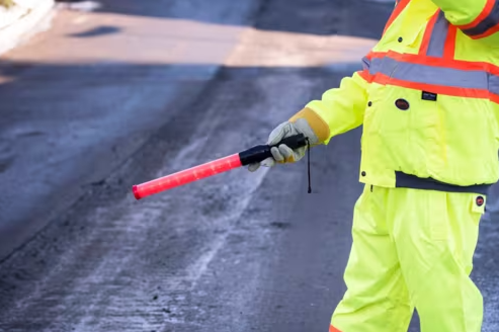 Person in neon yellow safety gear holding a red traffic baton, directing traffic on a road.