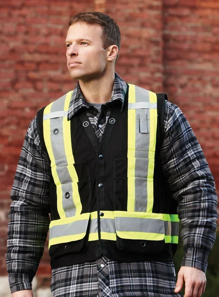 Man wearing a high-visibility vest over a plaid shirt, standing in front of a brick wall.