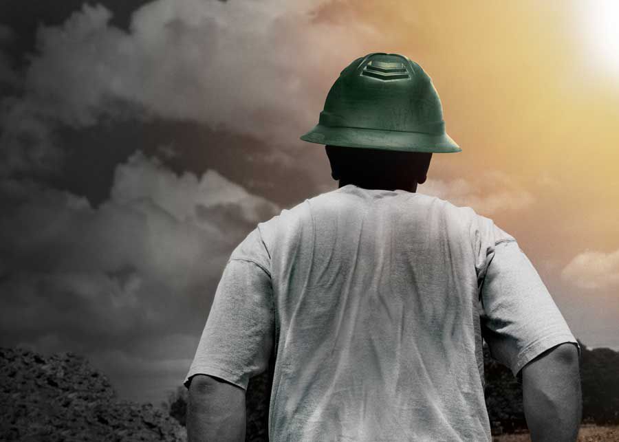 Person wearing green hard hat, facing bright sunlight breaking through dark clouds.