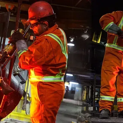 Two workers in orange protective suits and hard hats work with metal in a factory setting.