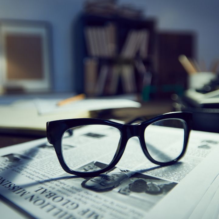 Black eyeglasses on a newspaper, shadows cast across a desk with books in the background. Private Investigator Services in Columbia TN 
