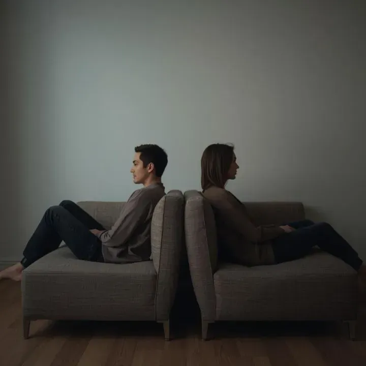 Two people sit back-to-back on separate sections of a sofa, facing away from each other.