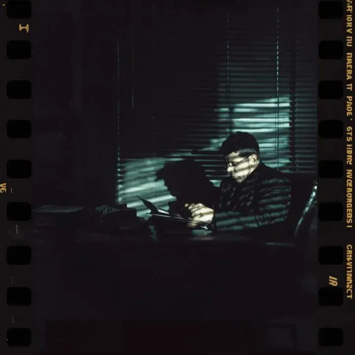 Man sitting at a desk, lit by light through blinds. Dark room, holding papers, reading.