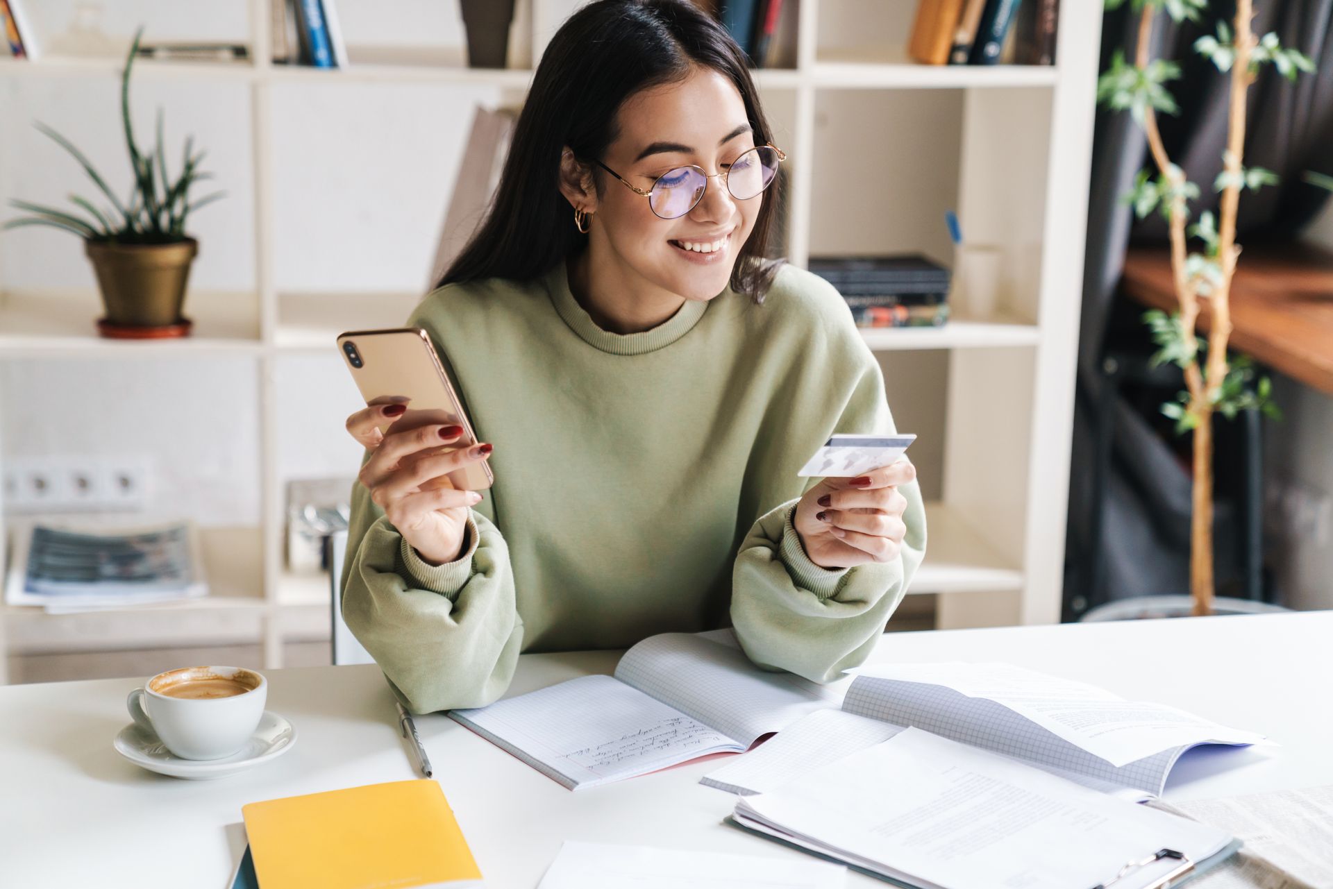 A woman is sitting at a table holding a credit card and a cell phone.