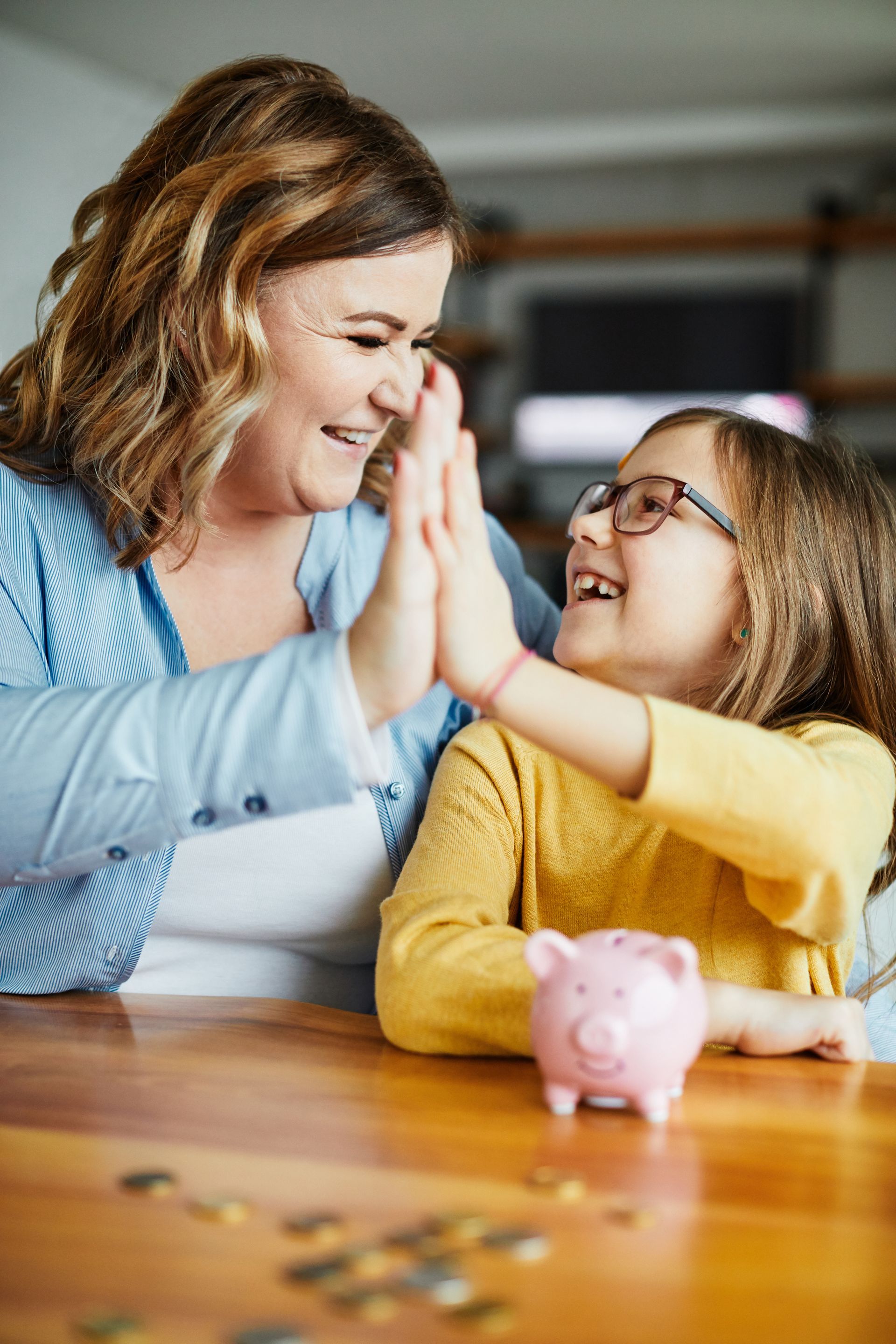 A woman and a little girl are giving each other a high five while sitting at a table with a piggy bank.