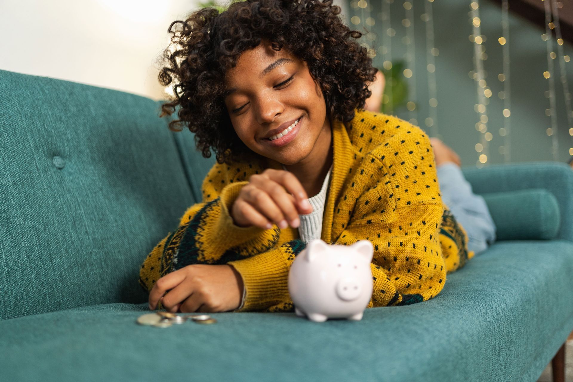A young girl is laying on a couch putting coins into a piggy bank.