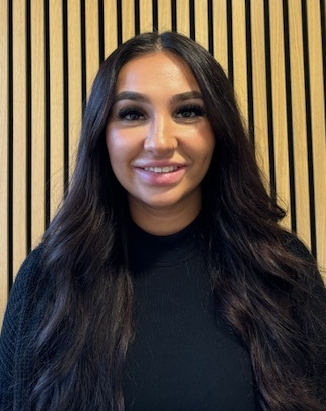 Woman with long dark hair, smiling in front of a wooden slatted wall, wearing a black top.