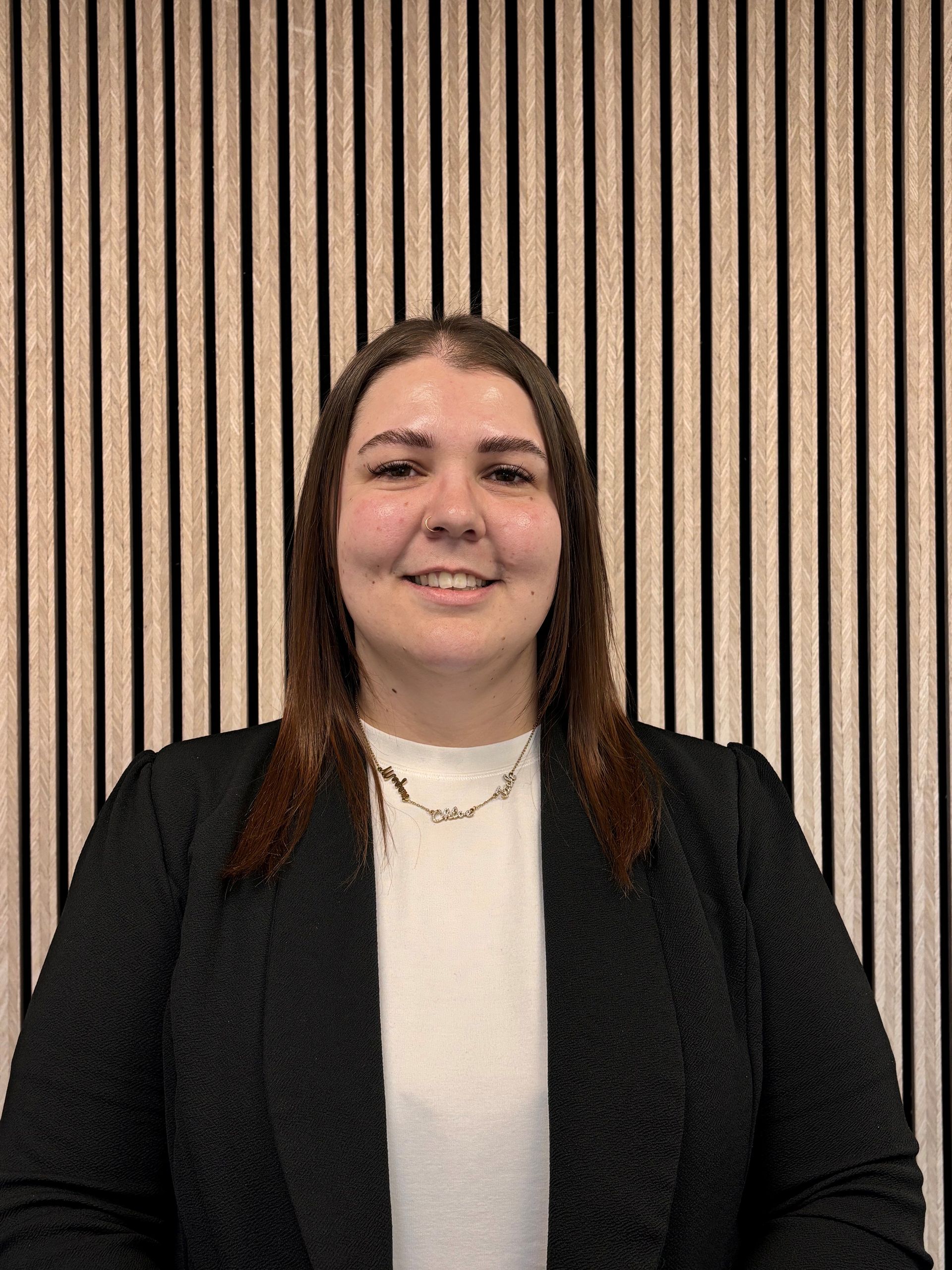 Woman in black blazer smiles at the camera, standing in front of a wooden panel wall.