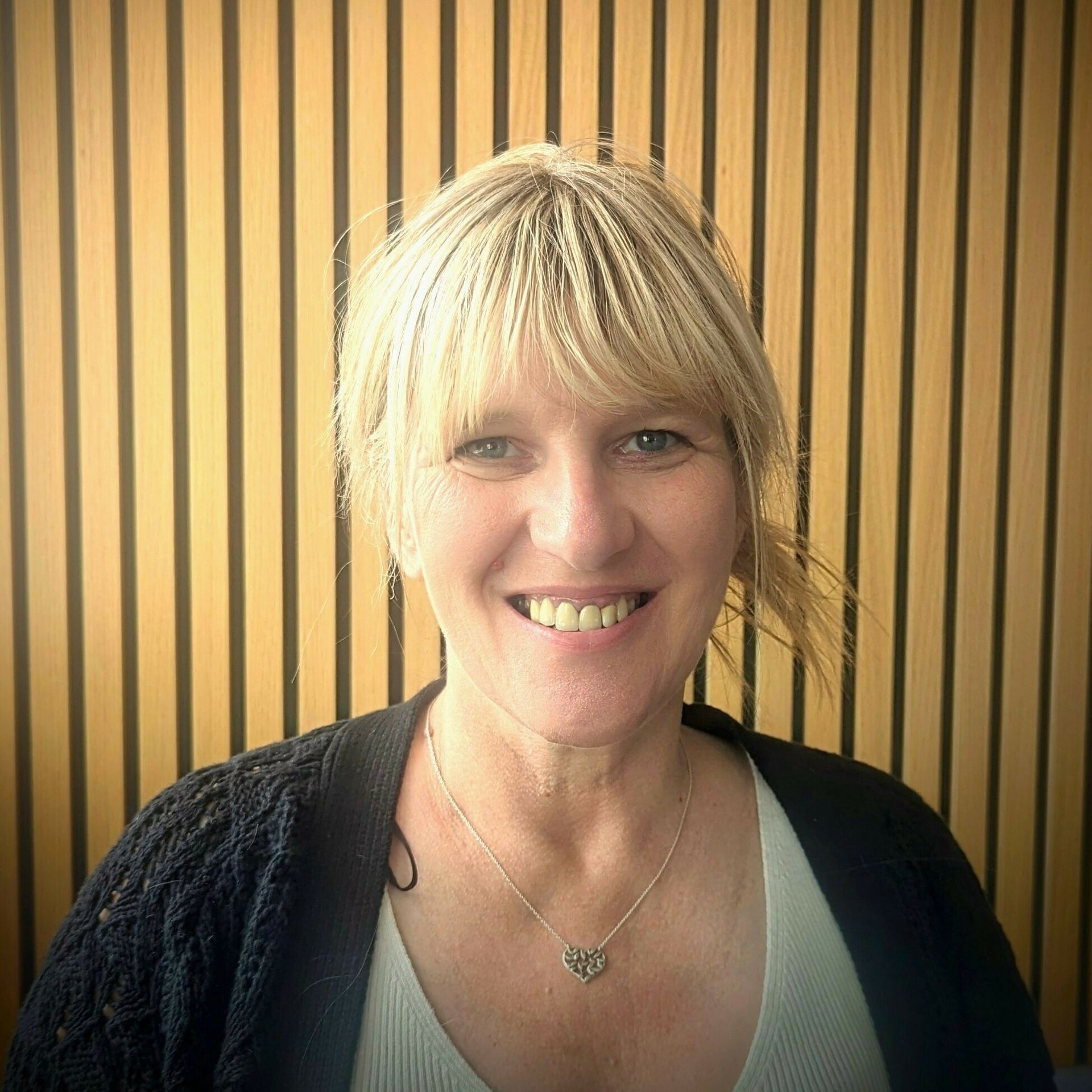 Woman with blonde hair smiling, wearing a black cardigan and necklace, in front of wood paneling.